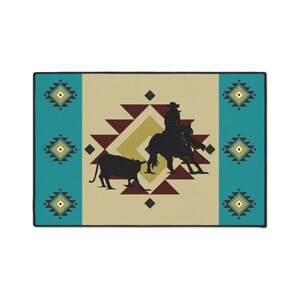 May include: A beige rug with a turquoise and maroon geometric pattern. A silhouette of a cowboy riding a horse and herding a cow is in the center of the rug.