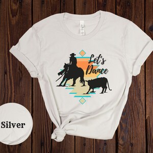 Op de afbeelding: Wit T-shirt met een silhouet van een cowgirl die een paard berijdt en een kalf. Het ontwerp bevat een turquoise en oranje geometrisch patroon en de tekst "Let's Dance".