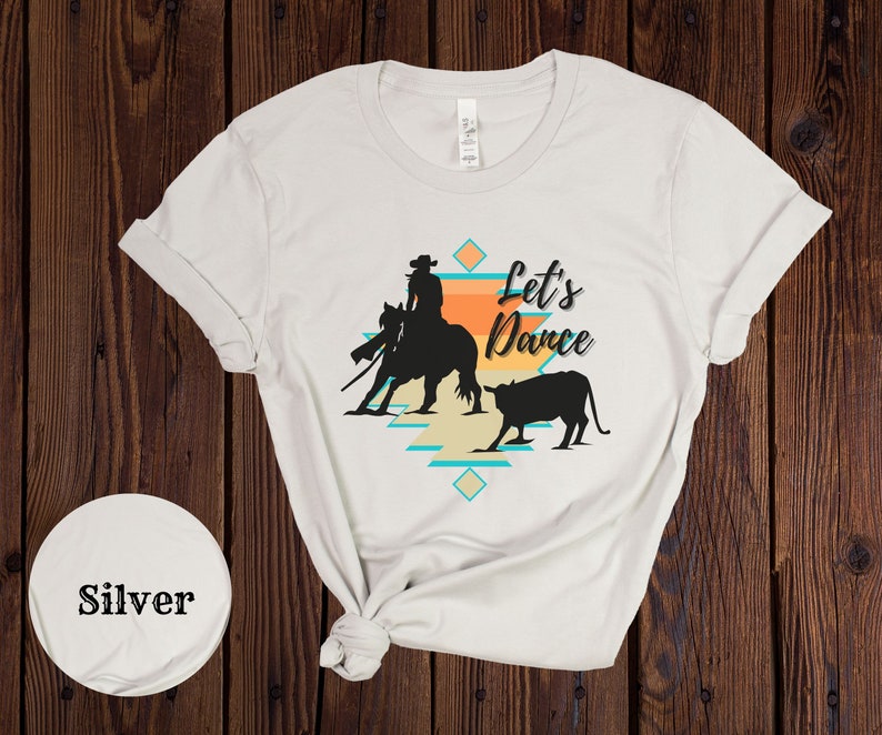 Op de afbeelding: Wit T-shirt met een silhouet van een cowgirl die een paard berijdt en een kalf. Het ontwerp bevat een turquoise en oranje geometrisch patroon en de tekst "Let's Dance".