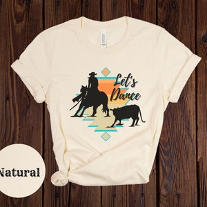 Op de afbeelding: Een cr&egrave;mekleurig T-shirt met een silhouet van een cowgirl die een paard berijdt en een kalf. De tekst "Let's Dance" is op het shirt gedrukt.