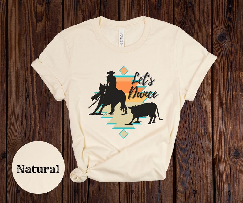 Op de afbeelding: Een cr&egrave;mekleurig T-shirt met een silhouet van een cowgirl die een paard berijdt en een kalf. De tekst "Let's Dance" is op het shirt gedrukt.