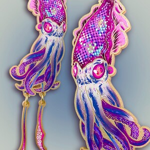 May include: Squid-shaped earrings in shades of purple, pink, and blue with gold accents. The earrings feature detailed designs on the squid's head and tentacles, with dangling gold chains. The background is a gradient of light blue and gray.