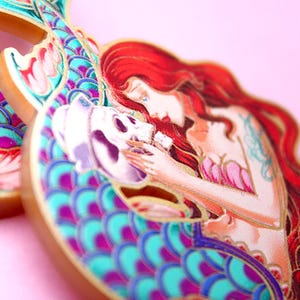 Könnte beinhalten: Eine farbenfrohe Emaille-Anstecknadel mit einer Meerjungfrau mit roten Haaren, die einen Schädel hält. Die Meerjungfrau trägt einen Muschelbh und hat einen türkisfarbenen und violetten Schwanz mit goldenen Akzenten.