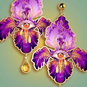May include: A pair of ornate earrings featuring a floral design with purple, gold, and orange hues. Each earring is shaped like a stylized iris flower, with an eye at the center and a teardrop-shaped charm.