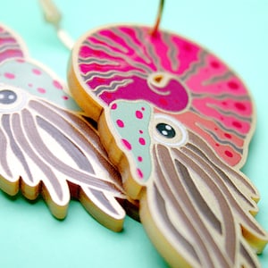 Op de afbeelding: Een paar gouden oorbellen met een nautilus-schelpontwerp in een roze, rode en groene kleurstelling. De nautilus-schelp is gedetailleerd met een spiraalvormig patroon en een klein oog.