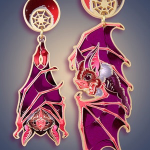 May include: A pair of earrings featuring a detailed illustration of bats with wings in a deep purple and pink color scheme. The bats are depicted with a web design and a red moon.