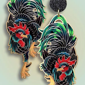 Könnte beinhalten: Ein Paar goldfarbene Ohrringe mit einem schwarzen und grünen Hahn-Design mit roten Akzenten. Die Ohrringe haben einen glitzernden schwarzen Anhänger oben.