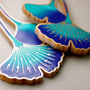 Könnte beinhalten: Nahaufnahme von zwei Ohrringen aus Holz in Form von Ginkgoblättern. Die Blätter sind mit einem Farbverlauf aus Blau und Türkis bemalt, mit goldenen Akzenten und weißen Konturen. Die Ränder der Ohrringe sind in Naturholzfarbe gehalten.