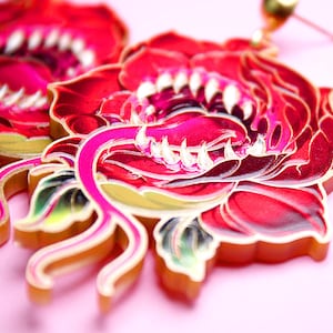 Könnte beinhalten: Nahaufnahme von roten und rosa Blumenohrringen mit weißen zahnartigen Details. Die Ohrringe haben ein mehrschichtiges Design mit goldenen Akzenten und einem rosa Hintergrund. Ein einzigartiges Accessoire.