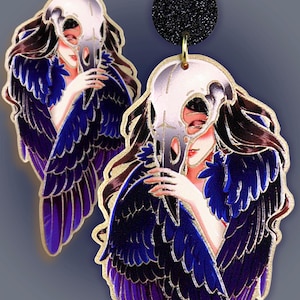 Könnte beinhalten: Künstlerische Ohrringe mit einer Frau mit langen braunen Haaren, die eine Totenkopfmaske hält. Die Ohrringe haben blaue und violette Federflügel mit goldenem Rand. Der obere Teil des Ohrrings ist ein schwarzer Kreis.