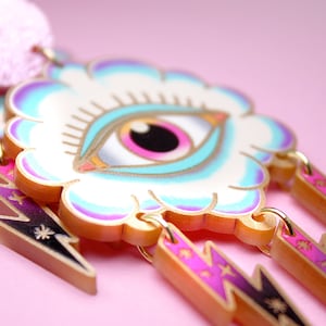 Può includere: Orecchini dorati con un ciondolo a forma di nuvola bianca che presenta un occhio blu e rosa con ciglia. Il ciondolo a forma di nuvola è attaccato a tre ciondoli a forma di fulmine dorati con accenti rosa e viola.