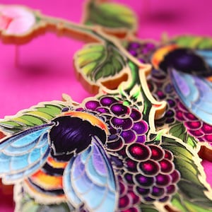 Pode incluir: Close-up de uma representação artística colorida de abelhas e amoras. O design apresenta uma abelha com asas azuis e amarelas e um cacho de amoras roxas. O fundo é rosa vibrante.