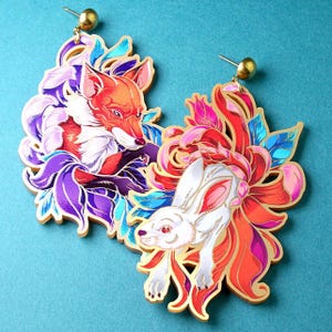 Könnte beinhalten: Ein Paar goldfarbene Ohrringe mit einem Fuchs und einem Hasen. Der Fuchs ist orange und weiß, umgeben von lila und blauen Akzenten. Der Hase ist weiß mit rosa Ohren, vor roten und orangefarbenen Blumendesigns. Die Ohrringe haben goldene Stecker.