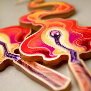 Op de afbeelding: Twee platte, decoratieve ornamenten in de vorm van lucifers met een levendig, wervelend vlamontwerp in tinten rood, oranje, geel en magenta. De luciferkoppen zijn paars met witte accenten en de stokjes hebben een gemarmerd, veelkleurig patroon.