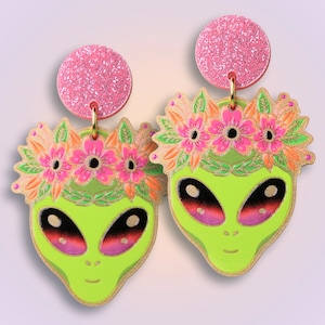 Könnte beinhalten: Ein Paar grüne Alien-Gesichtsohrringe mit rosa Glitzer-Blumenkronen. Die Ohrringe sind aus Holz und haben einen rosa Glitzer-Oberteil.