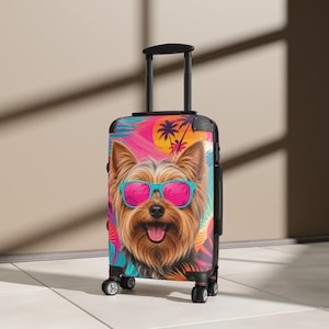 May include: A black rolling suitcase featuring a vibrant, colorful graphic of a Yorkshire Terrier wearing bright turquoise sunglasses. The background design includes palm trees and sunset-inspired hues of pink, orange, and yellow.