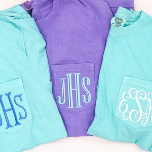 May include: Three t-shirts with embroidered monograms on the pocket. Two of the shirts are light blue and one is purple.