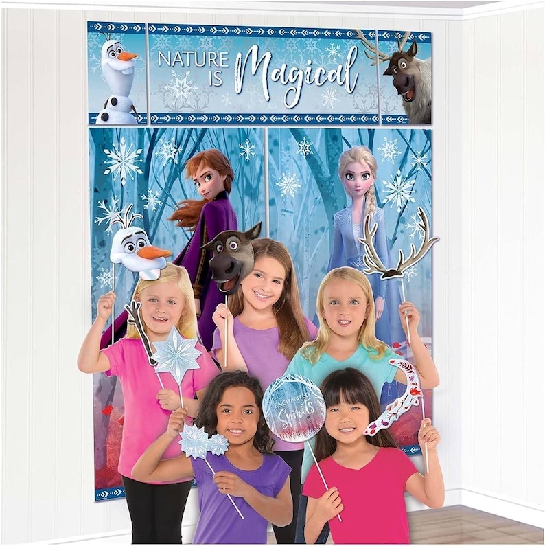May include: A group of children pose with character-themed photo booth props in front of a backdrop featuring characters from the movie Frozen. The backdrop reads "Nature is Magical". The children are wearing various colored shirts.