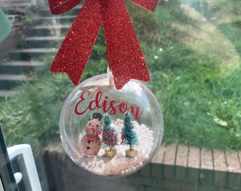 May include: A clear glass Christmas bauble with a red bow, a snowman, two small pine trees, and white beads inside. The bauble has the name "Edison 2020" written on it.