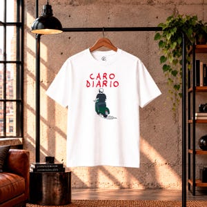 Puede incluir: Una camiseta blanca de manga corta con un gráfico de una persona montando un scooter verde. Sobre la ilustración, el texto "CARO DIARIO" está impreso en rojo. La camiseta tiene cuello redondo y se muestra en una percha de madera.