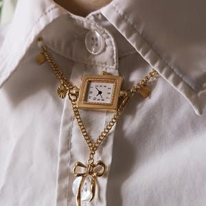 Puede incluir: Un collar dorado con un colgante de reloj cuadrado, esfera blanca y números negros. El collar incluye un dije en forma de lazo con un centro redondo y transparente, usado sobre una camisa blanca.