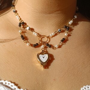 Puede incluir: Collar dorado con un colgante de reloj en forma de corazón. El collar tiene dos hilos de cuentas en colores negro, blanco y perla. La esfera del reloj es blanca con números y agujas negras. El collar se lleva alrededor del cuello.