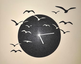 以下が含まれることがあります： 白い針と、周りを飛ぶ黒い鳥の群れがある黒い壁掛け時計。 時計の文字盤は、テクスチャ加工された黒い円です。 鳥はさまざまなサイズと位置にあります。