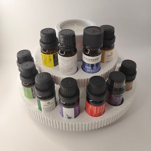 May include: A white ceramic tiered stand with 12 slots for essential oil bottles. The stand has a small white candle in the center. The bottles are various colors and sizes, with labels that include the words "Eucalyptus", "Time to Rest", and "Lemon".