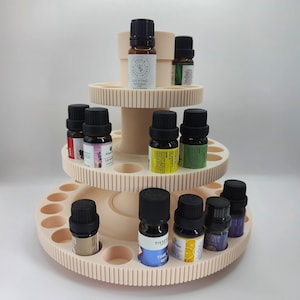 Könnte beinhalten: Ein dreistufiger, hellrosa ätherischer Öl-Präsentationsständer. Der Ständer hält mehrere kleine Glasflaschen mit schwarzen Kappen, die jeweils mit verschiedenen ätherischen Ölnamen beschriftet sind. Der Ständer hat ein zahnradartiges Design.