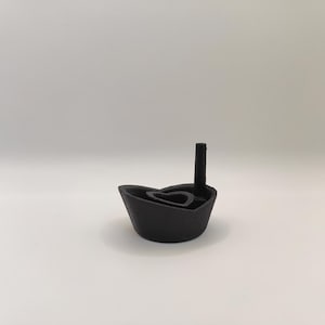 May include: A black, boat-shaped candle holder with a heart-shaped insert and a tall, cylindrical element. The candle holder is made of a matte material and sits on a white surface. The design is minimalist and modern.