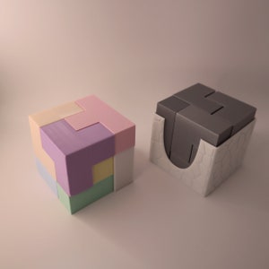 May include: Two 3D printed puzzles, one in pastel colours and one in grey, with a white base. The pastel puzzle is assembled into a cube shape, while the grey puzzle is disassembled and sitting on its base.