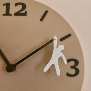 May include: A beige wall clock with black hands and numerals. A white figure is attached to the minute hand, appearing to hang from it. The clock face has the numbers 12 and 3 clearly visible.