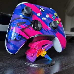 Puede incluir: Un mando de Xbox azul y rosa con un diseño personalizado se muestra en un soporte con un logotipo impreso en 3D. El logotipo es un diseño "8 bits" estilizado con un esquema de colores arcoíris.