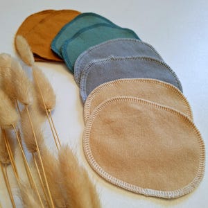 Könnte beinhalten: Ein Set mit sieben wiederverwendbaren Abschminkpads in verschiedenen Farben, darunter Senfgelb, Teal, Grau und Beige. Die Pads sind aus weichem Stoff und haben einen genähten Rand.