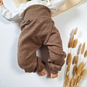 Könnte beinhalten: Eine braune Hose mit hoher Taille, breitem, umgeschlagenem Bund und Bündchen an den Knöcheln, getragen über einem weißen Langarmshirt. Die Hose besteht aus strukturiertem Stoff und hat ein kleines, dunkles, rechteckiges Etikett an der Seite.
