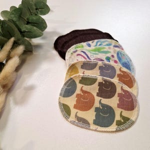 Könnte beinhalten: Ein Stapel wiederverwendbarer, oval geformter Abschminkpads. Die Pads weisen verschiedene Muster auf, darunter ein braunes Rippgewebe, ein blau-weißes Blumenmuster und ein beiges Gewebe mit bunten Elefantenmotiven. Die Ränder sind mit weißen Nähten versehen.