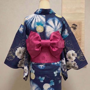 May include: A navy blue yukata, a traditional Japanese garment, is displayed on a mannequin. The yukata features a vibrant pink obi sash tied in a bow. The fabric is patterned with white, blue, and yellow floral and abstract designs.