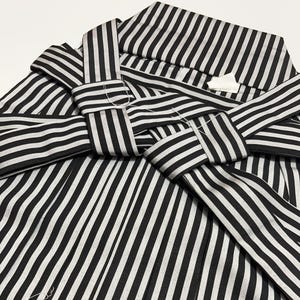 May include: Close-up of a black and white striped garment, possibly a kimono or robe. The fabric is folded and tied, creating a layered effect. The stripes are vertical and evenly spaced, with a contrasting white background. White thread is visible.