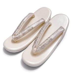 May include: A pair of off-white geta sandals. The sandals have a smooth, rounded base and feature silver and white floral patterned straps. The sandals are designed for traditional Japanese attire.