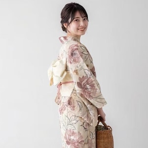 May include: A woman wearing a floral patterned kimono with a butterfly knot obi. The kimono features large mauve and cream-colored flowers and green leaves. She is holding a woven basket and wearing wooden sandals.
