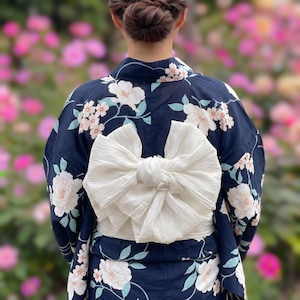 May include: A navy blue yukata, a traditional Japanese garment, with white floral patterns and a large white obi bow. The floral design includes white and light blue accents. The background is a soft focus of pink flowers.