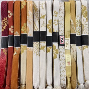 May include: A collection of colorful, patterned fabric belts. The belts vary in color, including red, orange, gold, and white, with floral and geometric designs. Each belt is held together with a black band.