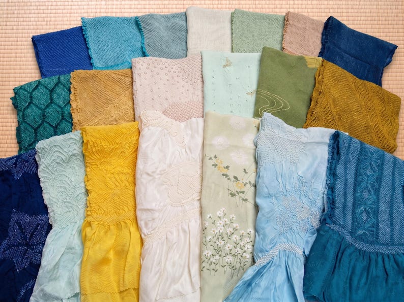 May include: A collection of colorful, textured fabrics in shades of blue, green, yellow, and white. Some fabrics feature intricate patterns and floral designs. The textiles are arranged on a woven mat, showcasing a variety of textures and hues.