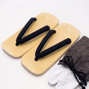 May include: A pair of light tan zori sandals with black fabric thong straps and a textured sunburst pattern on the footbed, accompanied by a pair of white split-toe tabi socks tucked inside a dark gray drawstring fabric storage pouch.