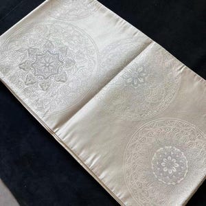 May include: A rectangular, off-white pillow with a detailed, circular, floral pattern. The pillow is made of a smooth, satin-like material and is placed on a dark, textured surface.