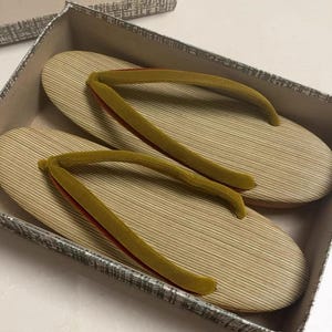 May include: A pair of traditional Japanese geta sandals in a box. The sandals have a light brown, striped base with olive green straps and red accents. The box is gray with a patterned design.