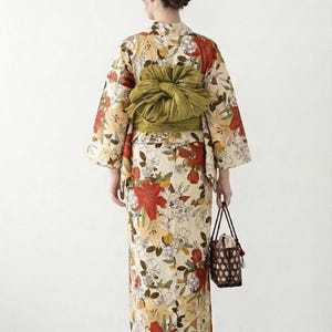 K&ouml;nnte beinhalten: Blumenkimono mit cremefarbenem Hintergrund, mit roten, gelben und wei&szlig;en Blumen. Der Kimono ist mit einem gr&uuml;nen Obi gebunden. Eine kleine, braune, gewebte Handtasche wird in der Hand gehalten.