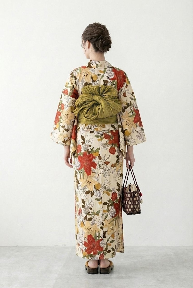 K&ouml;nnte beinhalten: Blumenkimono mit cremefarbenem Hintergrund, mit roten, gelben und wei&szlig;en Blumen. Der Kimono ist mit einem gr&uuml;nen Obi gebunden. Eine kleine, braune, gewebte Handtasche wird in der Hand gehalten.