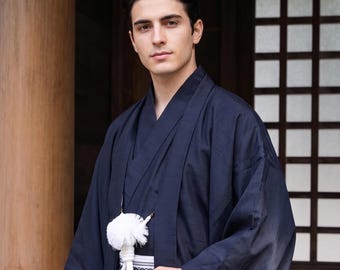 May include: A dark navy blue kimono with wide sleeves and a structured collar, worn over a patterned obi sash. A white, fluffy, circular decorative accessory hangs from the front of the garment by a thick white cord, resting against the dark fabric.