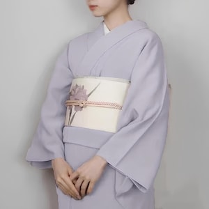 May include: A woman wearing a light purple kimono with a white obi featuring a floral design. The obi is tied with a decorative knot.